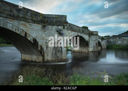 Scenic view of bridge spanning River Wharfe flowing gently through Burnsall village, on summer evening with blue sky - Yorkshire Dales, England, UK. Stock Photo