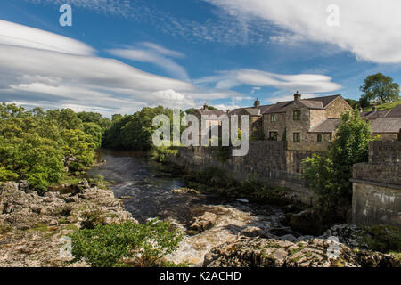 Blue sky over cascading water & riverside houses at sunny, scenic Linton Falls waterfall over River Wharfe, Grassington, Yorkshire Dales, England, UK. Stock Photo