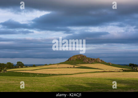 Under dark clouds & across multi-coloured fields, view of distinctive high rocky outcrop of Almscliffe Crag lit by sun - North Yorkshire, England, UK.
