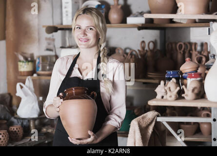 Cheerful woman in apron carrying ceramic vessels in atelier Stock Photo