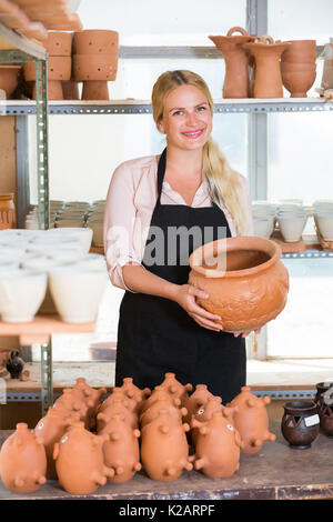 Glad woman potter in apron carrying ceramic vessels in atelier Stock Photo