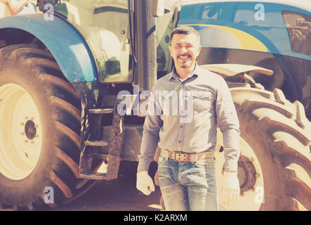 portrait of positive smiling mature male farmer near huge tractor Stock Photo