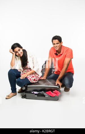 stock photo of indian young Couple packing for holiday, trying to close suitcase full with clothes, sitting isolated over white background Stock Photo