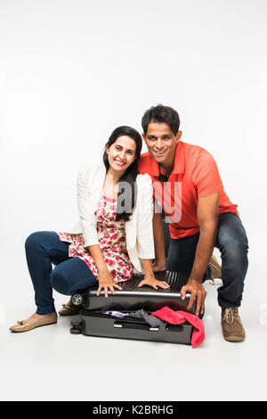 stock photo of indian young Couple packing for holiday, trying to close suitcase full with clothes, sitting isolated over white background