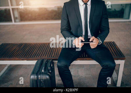 Cropped shot of young businessman sitting on bench with suitcase and using digital tablet. Man waiting in public transport station. Stock Photo