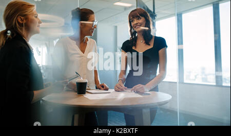 Three female executives standing and discussing around a table. Corporate professional having an informal meeting in modern office. Stock Photo