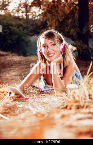Cute smiling young woman lying on the withered grasses and listening music. Looking at camera. Stock Photo
