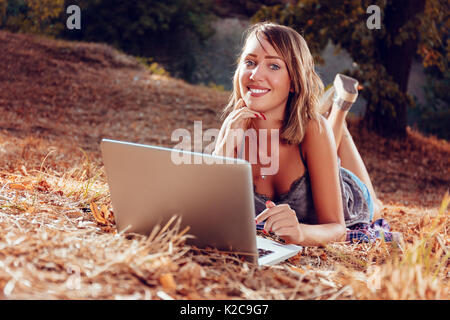 Cute smiling young woman lying on the withered grasses in autumn and using laptop. Stock Photo