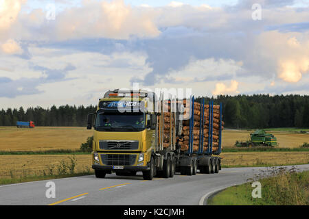 SALO, FINLAND - APRIL 21, 2017: Yellow Volvo FH16 logging truck transports a load of pine logs along country road in the evening with agricultural equ Stock Photo