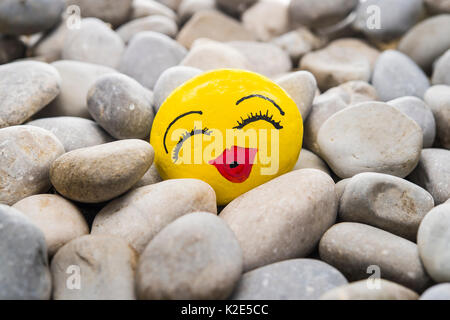 Smiley face made of stone, painted yellow with red kissing lips, lies in between stones Stock Photo