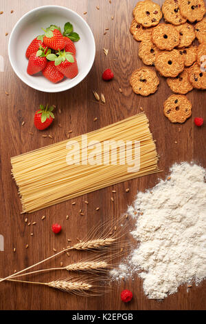 Top view of dry spaghetti pasta, flour, wheat ears, homemade cookies, strawberries in a white ceramic bowl and raspberries on dark wooden table Stock Photo