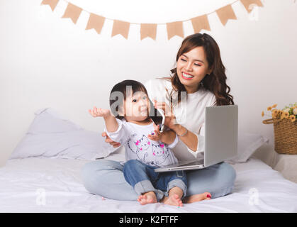 Asian lady in classic suitvworking on laptop at home with her baby girl chatting with father. Stock Photo
