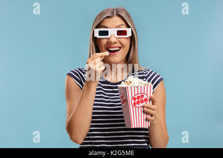 Woman wearing a pair of 3D glasses and eating popcorn isolated on blue background Stock Photo