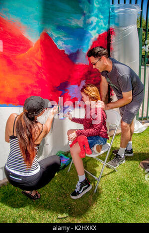 A young woman artist shows a girl and her supportive father some artistic technique as she paints a large outdoor abstract mural mounted on a fence in a Costa Mesa, CA, park during a weekend community festival. Stock Photo