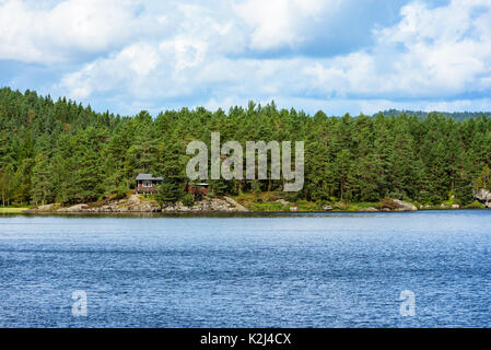 Small brown wooden cabin among the trees in the forest. Calm forest lake or river in foreground. Stock Photo