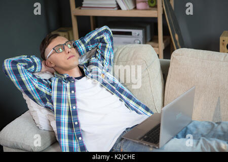 Young man working remotely lying on sofa. Hipster freelancer thinking while llying on couch with laptop. Stock Photo