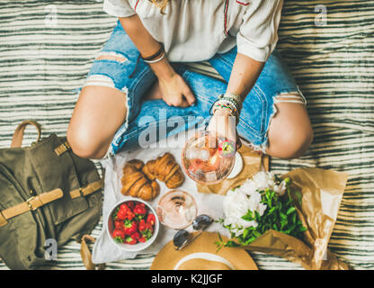 French style romantic picnic setting. Woman in jeans with glass of rose wine, fresh strawberries, croissants, brie cheese, sunglasses, peony flowers o Stock Photo