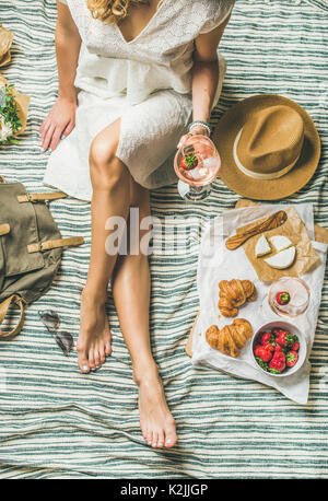 French style romantic picnic setting. Woman in dress with glass of wine, strawberries, croissants, brie cheese, sunglasses, straw hat, peony flowers o Stock Photo