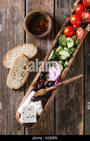 Ingredients for traditional greek salad. Cherry tomatoes, sliced cucumbers, red onion, black olives, feta cheese in olive wood bowl with loaf bread, o Stock Photo