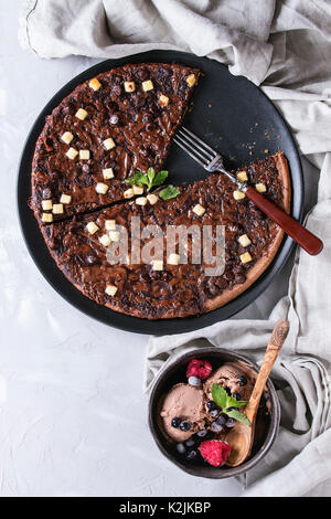 Dessert chocolate pizza with dark, milk, white chocolate, served on black plate with fork, mint, frozen berries and bowl of ice cream over gray concre Stock Photo