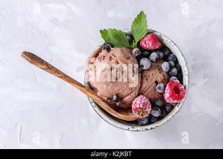 Homemade chocolate ice cream with frozen berries blueberry, raspberry, mint served in white bowl with olive wood spoon over gray concrete background. Stock Photo