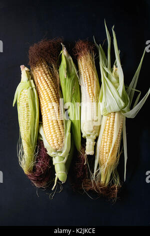 Young raw uncooked corn cobs in leaves. Top view over dark metal background. Stock Photo