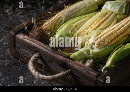 Young raw uncooked corn cobs in leaves in wooden tray. Close up over dark brown concrete texture background. Stock Photo