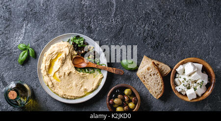 Homemade traditional spread hummus with chopping olives and herbs on blue plate, served with bread, olives, feta cheese, olive oil, spoon on black tex Stock Photo