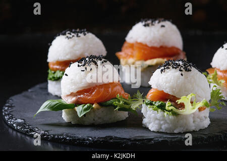 Mini rice sushi burgers with smoked salmon, green salad and sauces, black sesame served on slate stone board over black background. Modern healthy foo Stock Photo
