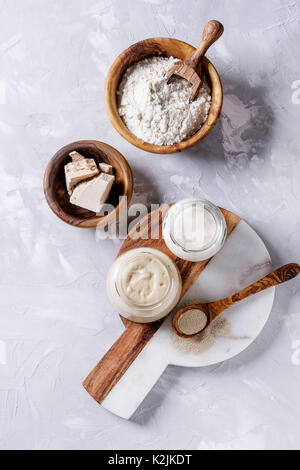 Rye and wheat sourdough in glass jars, fresh and instant yeast, olive wood bowl of flour for baking homemade bread. With spoon, serving board over gra Stock Photo