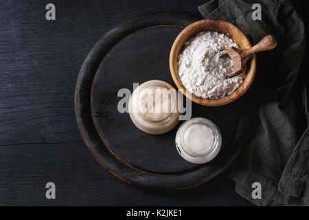 Rye and wheat sourdough in glass jars, olive wood bowl of flour for baking homemade bread. With scoop, serving board, textile over black burned wooden Stock Photo