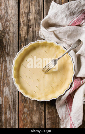 Dough for baking quiche tart in ceramic baking form ready for bake on kitchen towel over old plank wooden background. Flat lay with copy space Stock Photo