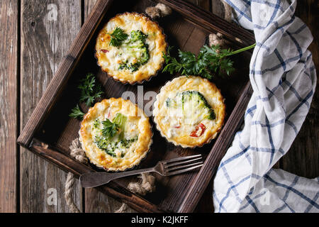 Baked homemade quiche pie in mini metal forms served with fresh greens, kitchen towel and fork in dark wood tray on old plank wooden background. Flat Stock Photo