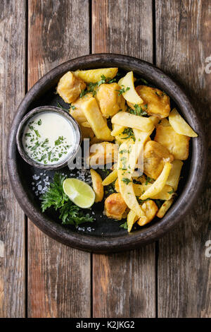 Traditional british fast food fish and chips. Served with white cheese sauce, lime, parsley, french fries in frying basket in terracotta tray over old Stock Photo