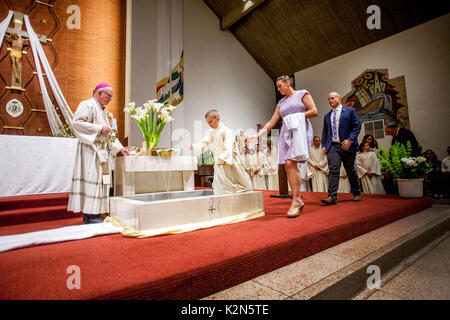 Accompanied by his parents and sponsors, a boy wearing a baptism robe enters the font at a Catholic cathedral in Orange, CA. Note bishop at left. Note other baptism candidates in background. Stock Photo