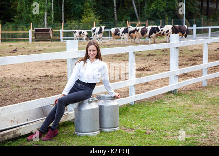 Cute young adult girl farmer, sitting on can after work. Two big can with milk. Pause after work in milk farm. Outdoor Stock Photo