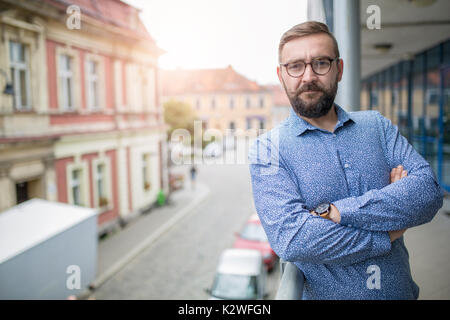 Elegant bearded fashion man standing at balcony with arms crossed and smiling Stock Photo