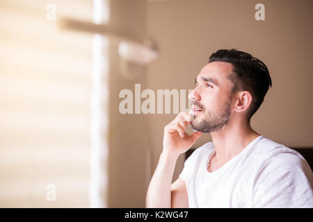 Young bearded latino talking on the phone in sunshine pouring through open window of his bedroom. Copy space on left side. Warm colors. Stock Photo