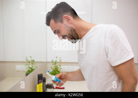 Joyful young man smiling during morning teeth brushing. He has black eyes and a beard. He is wearing white t-shirt. Stock Photo