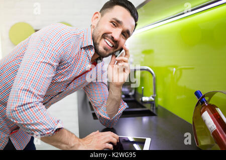 Smiling handsome man having nice conversation on the phone. He is standing in kitchen at kitchen counter and surfing the net on tablet. Stock Photo
