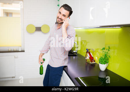 Man standing in kitchen and having phone conversation. He is holding two bottles of beer and smiling. Stock Photo