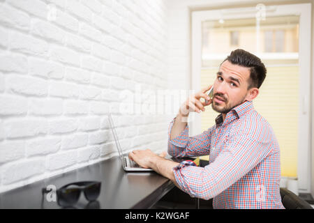 Young businessman talking on the phone in his kitchen. He is sitting at the table with laptop. Stock Photo
