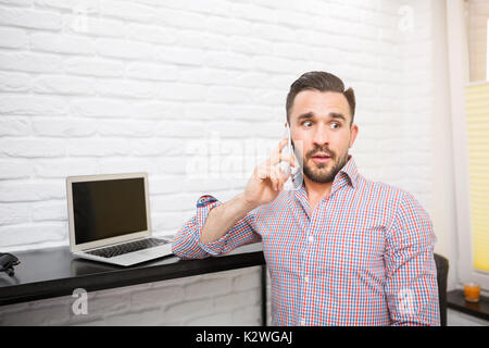 Surprised young man receiving some shocking news during phone call. He is working in kitchen on his laptop Stock Photo