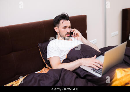 Man lying in bed with laptop and talking on the phone Stock Photo