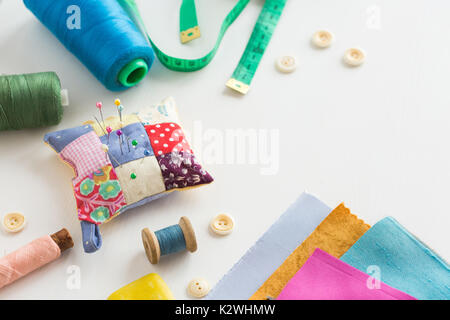 needlework, craft, sewing and tailoring concept - tools closeup on white desk, measuring meter, blue, pink and green threads in spools, scraps of colorful fabric, pincushion, white buttons and soap. Stock Photo