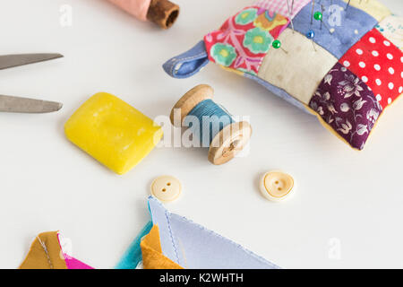sewing, patchwork, tailoring and fashion concept - closeup tools on white desk in studio, pincushion, blue and pink threads spools, white buttons, scissors, soap, pieces of colored patchwork fabric. Stock Photo