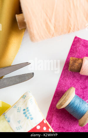 sewing, patchwork, tailoring and fashion concept - closeup tools on white work desk in studio, scissors, spools of blue and pink threads, pincushion, pieces of colored patchwork fabric, vertical. Stock Photo