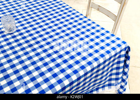 Empty table covered with blue and white chequered tablecloth next to white wooden chair. Stock Photo