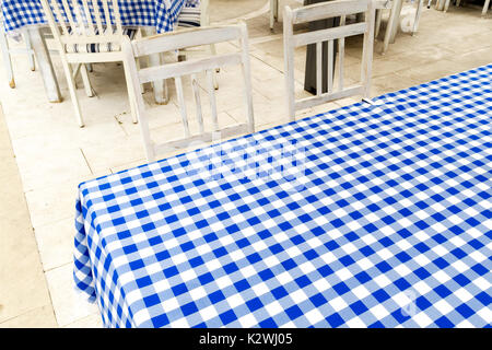 Empty table covered with blue and white chequered tablecloth next to white wooden chairs. Stock Photo
