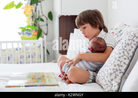 Sweet preschool boy, reading a book to his newborn brother, sitting on the bed in bedroom, childhood concept Stock Photo